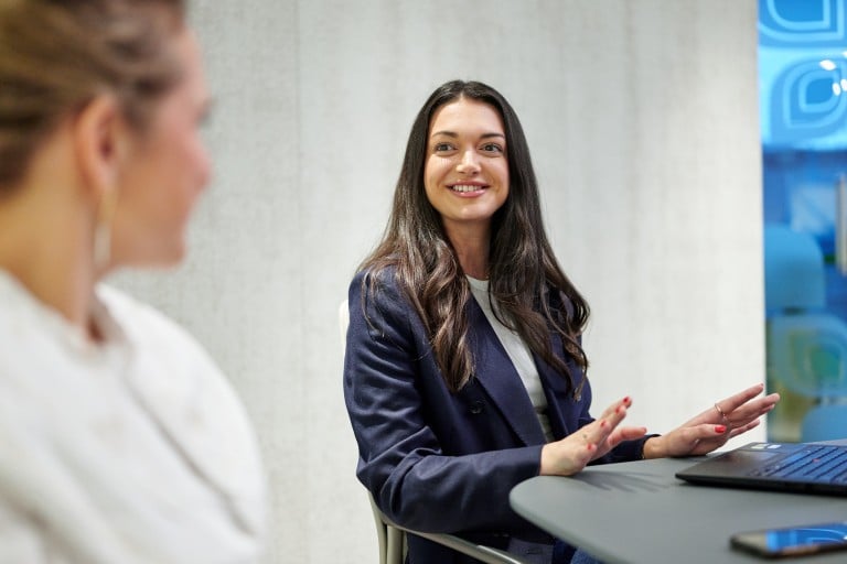 Eine Frau sitzt an einem Tisch in einem Besprechungsraum, lächelt und spricht gestikulierend mit einer anderen Person; vor ihr liegt ein Laptop.