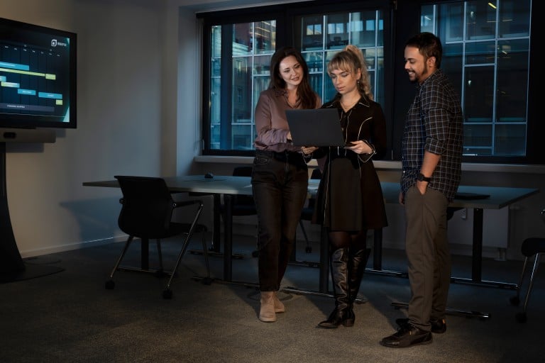 Drei Mitarbeitende stehen in einem modernen Büro zusammen und betrachten gemeinsam einen Laptop mit Projektinformationen auf dem Bildschirm im Hintergrund.