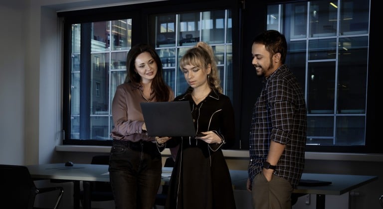 Three professionals collaborate over a laptop in a modern office, with a cityscape visible through large windows. The scene highlights teamwork, focus, and technology in a professional setting.