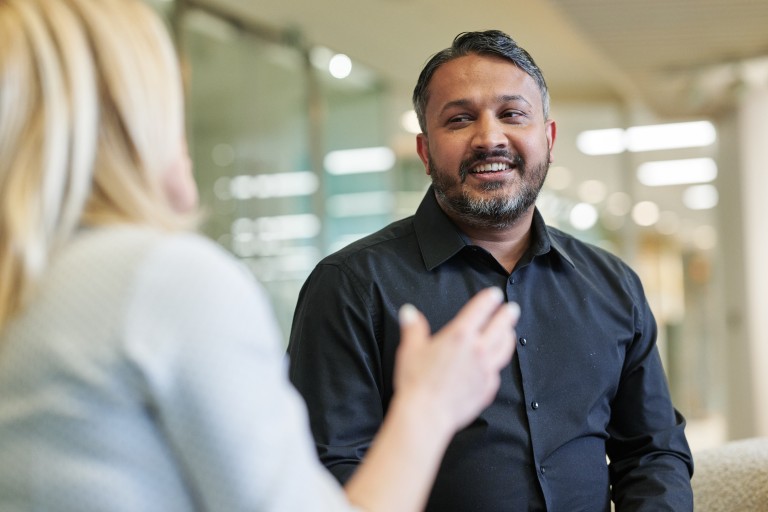 A man sits in an office, smiling as he talks with another person who is blurred in the foreground.