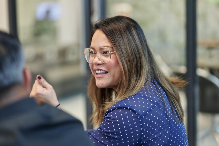 A person is seated indoors and engaged in conversation with another person in the foreground. The individual is wearing a patterned top and glasses. The background shows a blurred office area with glass partitions.