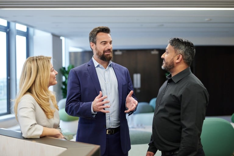 Three individuals are standing together in a modern indoor setting engaged in conversation. One individual is leaning on a piece of furniture, while the other two stand facing each other. Large windows and a bright, open office space are visible in the background.