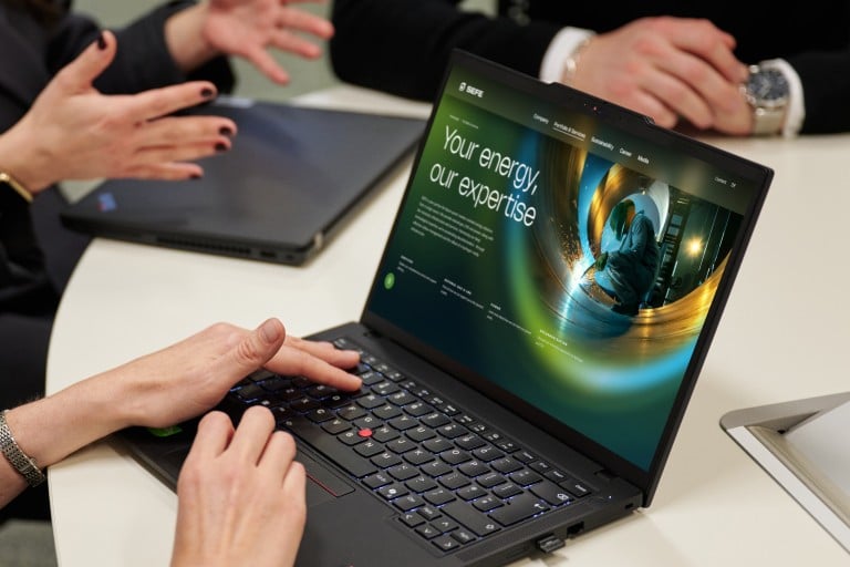 Several individuals are seated around a round table in a meeting room, working with laptops. One open laptop displays a website with the text ‘Your energy. Our expertise.’. Hand gestures indicate an ongoing discussion.
