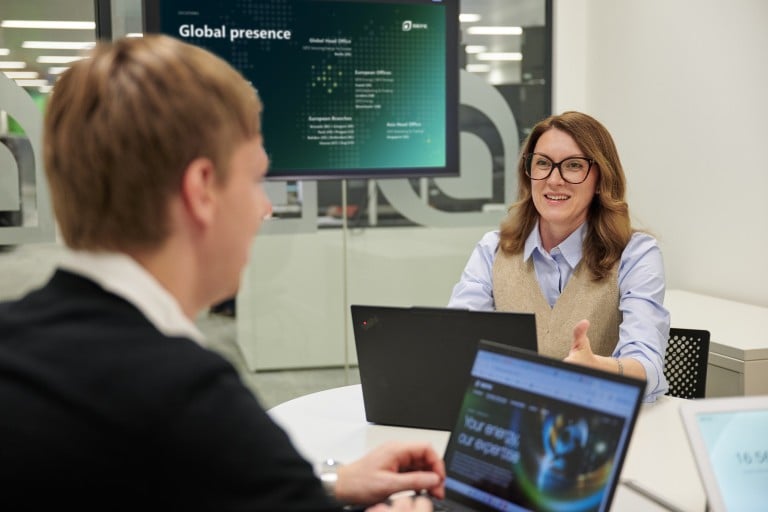 Two individuals are seated at a table in a meeting room, each working on a laptop. One individual gestures towards the other. In the background, a screen displays information about global locations and international presence.