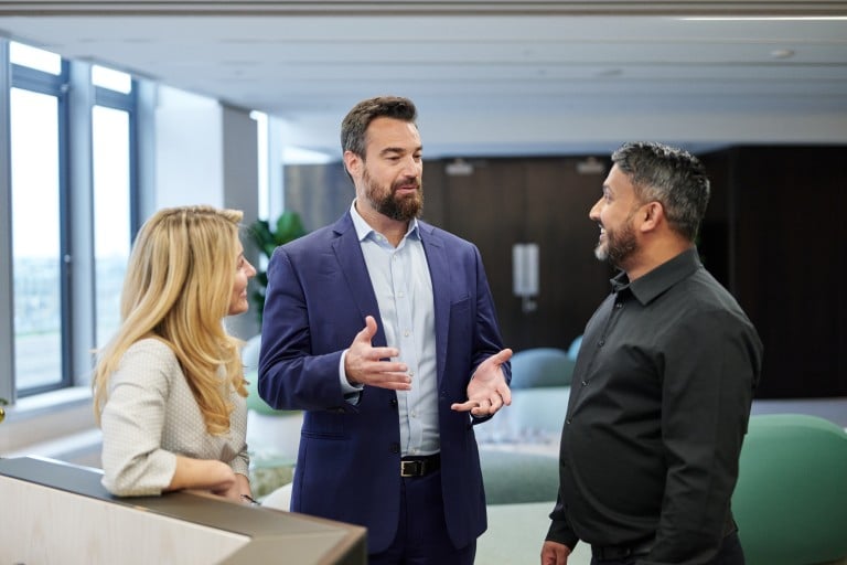 Three individuals are standing together in a modern indoor setting engaged in conversation. One individual is leaning on a piece of furniture, while the other two stand facing each other. Large windows and a bright, open office space are visible in the background.