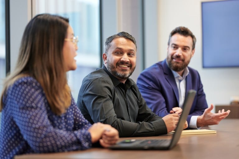 Three individuals are seated at a table in a meeting room. One individual in the foreground is shown from the side, while two others sit on the opposite side of the table. A laptop is placed in front of them. Large windows and a bright interior space are visible in the background.