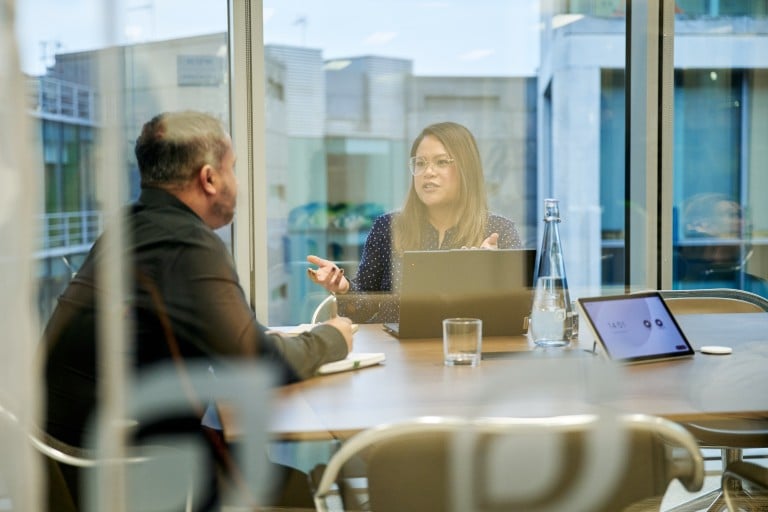Two people sit at a table in a bright meeting room; a woman gestures while explaining something as the other person listens. A laptop, tablet, water bottle and glass are on the table.