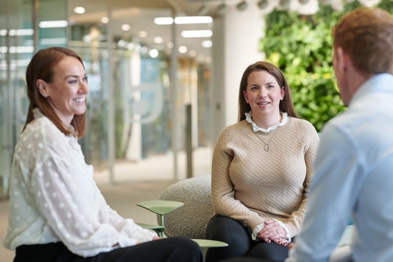 Three individuals are seated together in an open office area. Two individuals sit side by side on lounge chairs and look toward a third individual sitting opposite them. The background includes glass partitions, greenery and a modern interior setting.
